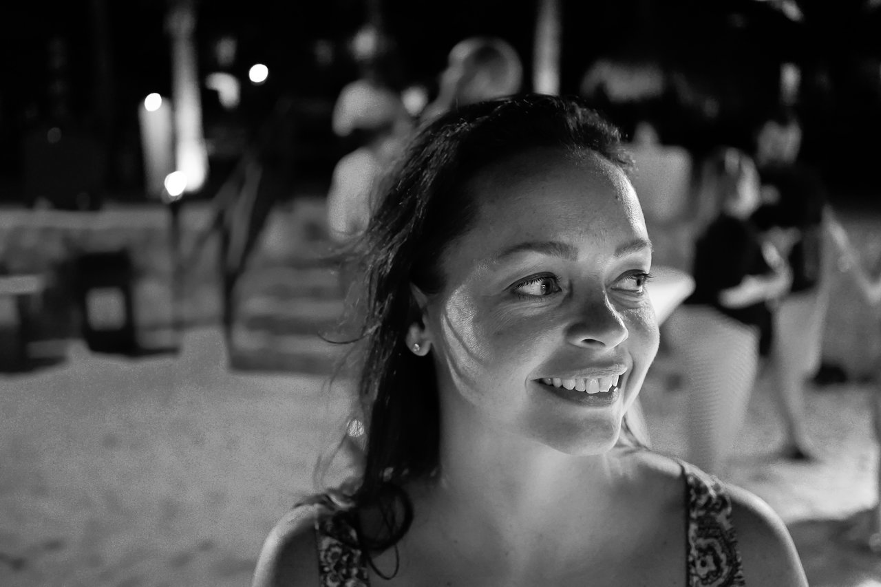 A woman smiles and looks to the side at an outdoor nighttime gathering with people in the background.