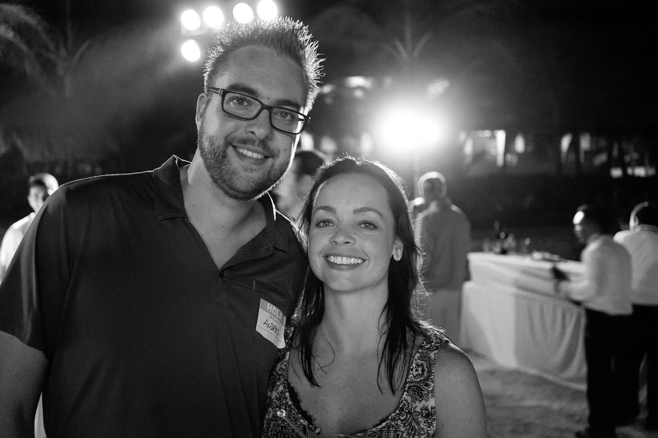 A man and woman smile at the camera at an outdoor nighttime event with people in the background.