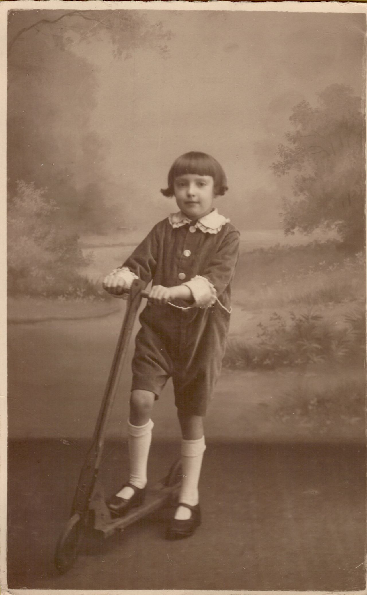 A young child stands on a wooden scooter, holding the handlebars, wearing a collared outfit with knee-high socks.
