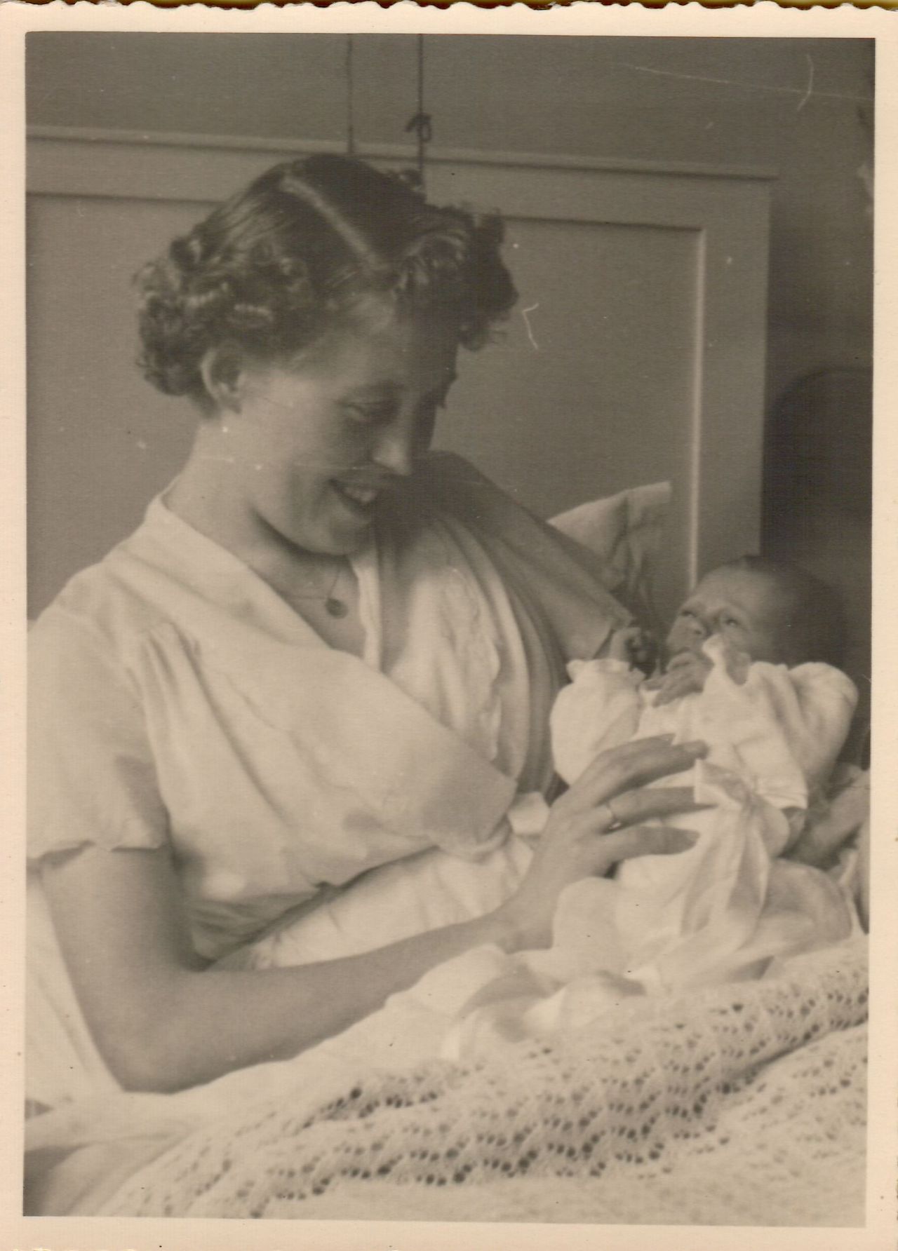 A woman smiles while holding a newborn baby dressed in white, gently cradling them in her arms.