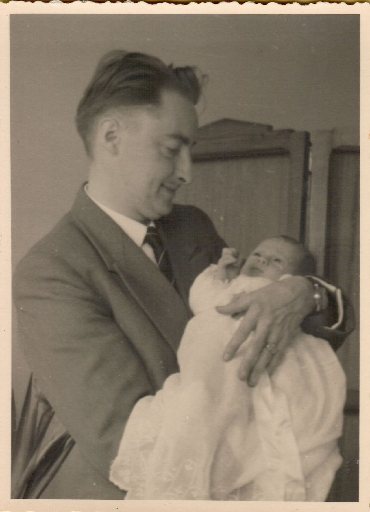 A man in a suit holds a newborn baby dressed in white, looking down with a gentle expression.