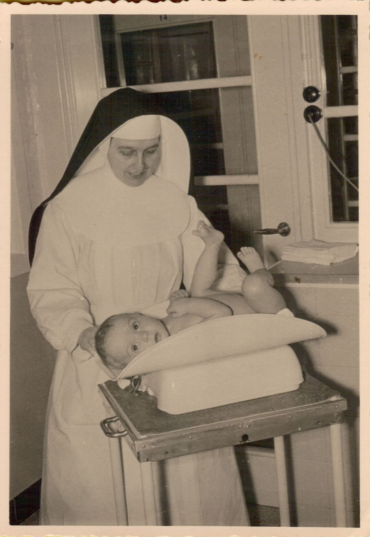 A nun gently holds a baby while weighing them on a scale in a room with a window.
