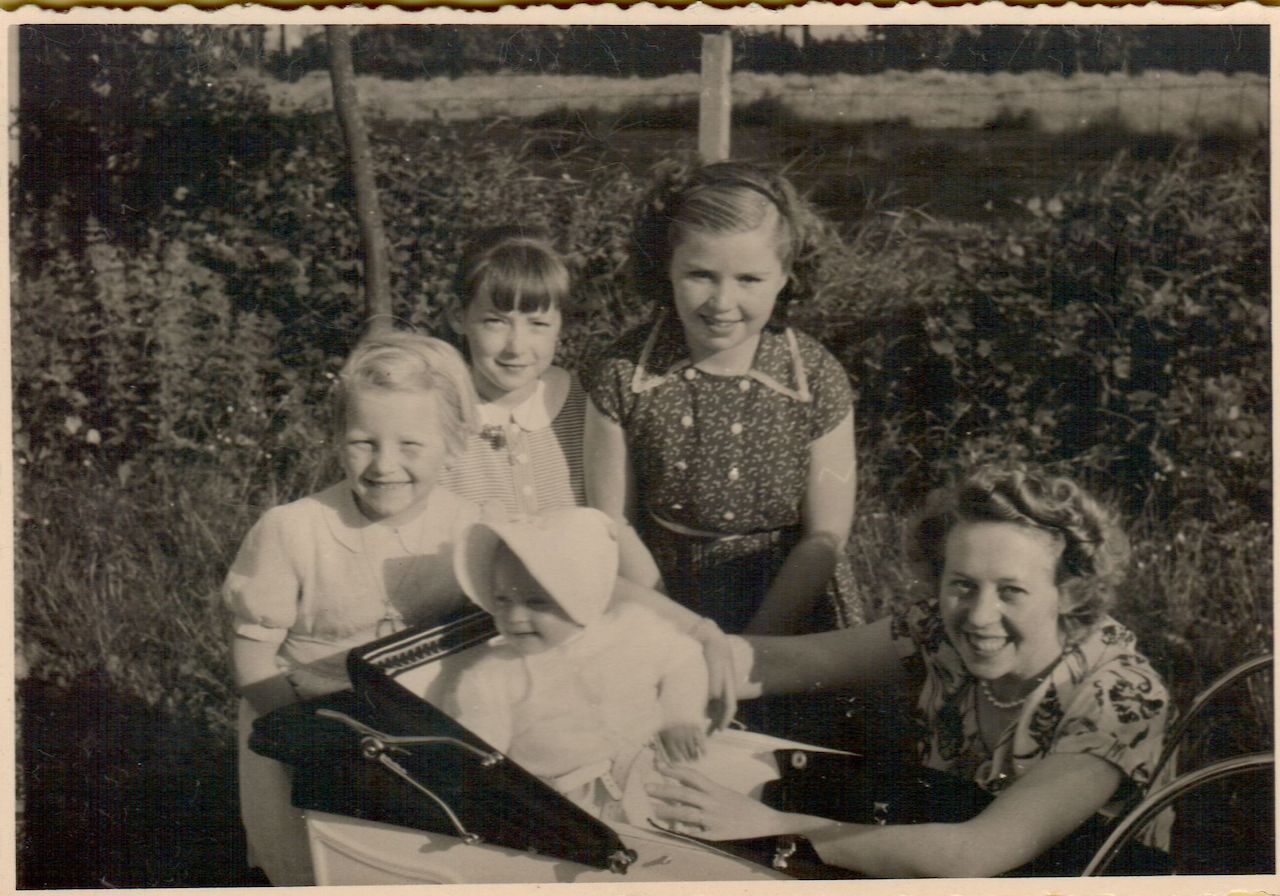 A smiling woman leans toward a baby in a stroller, surrounded by three young girls outdoors.