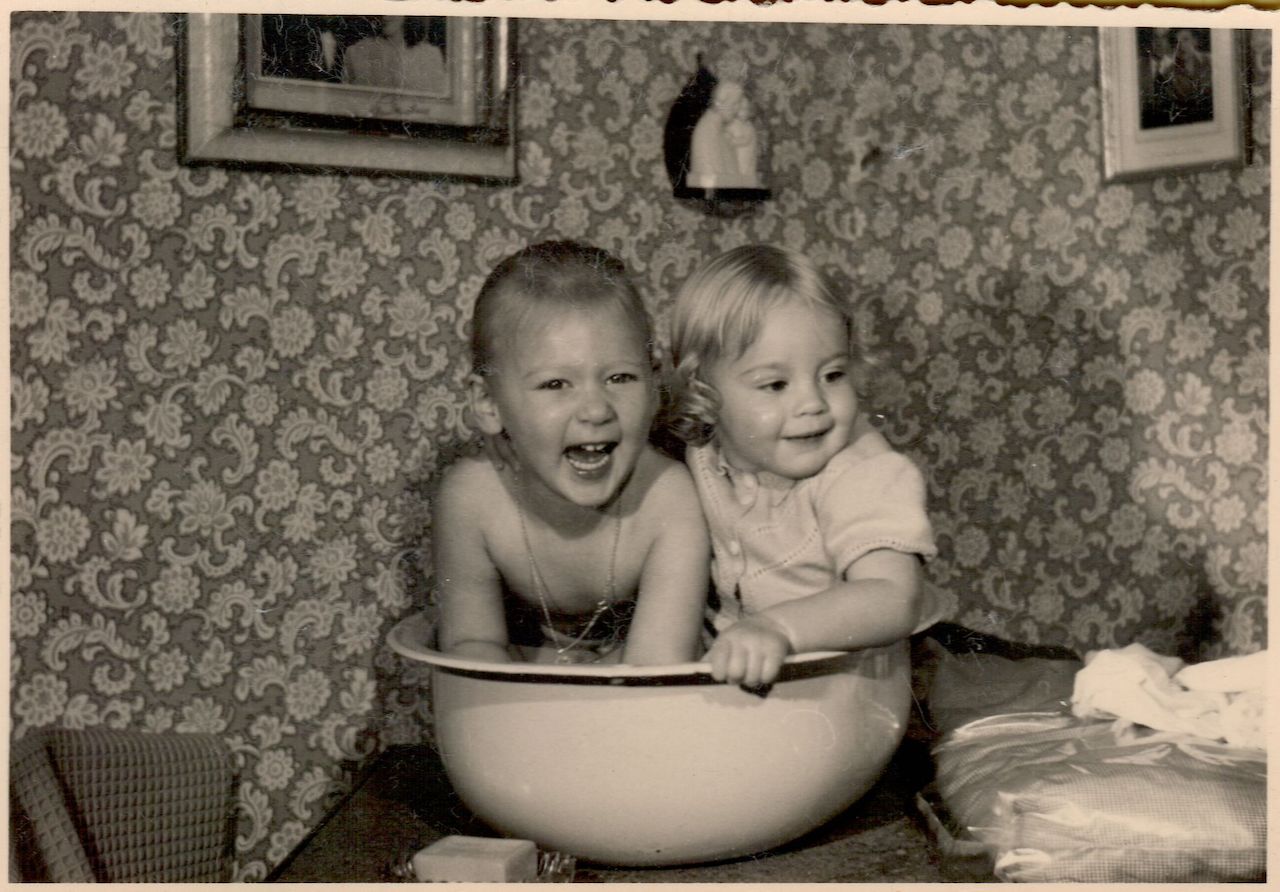 Two young children sit together in a large basin, one laughing and the other smiling, in a vintage indoor setting.