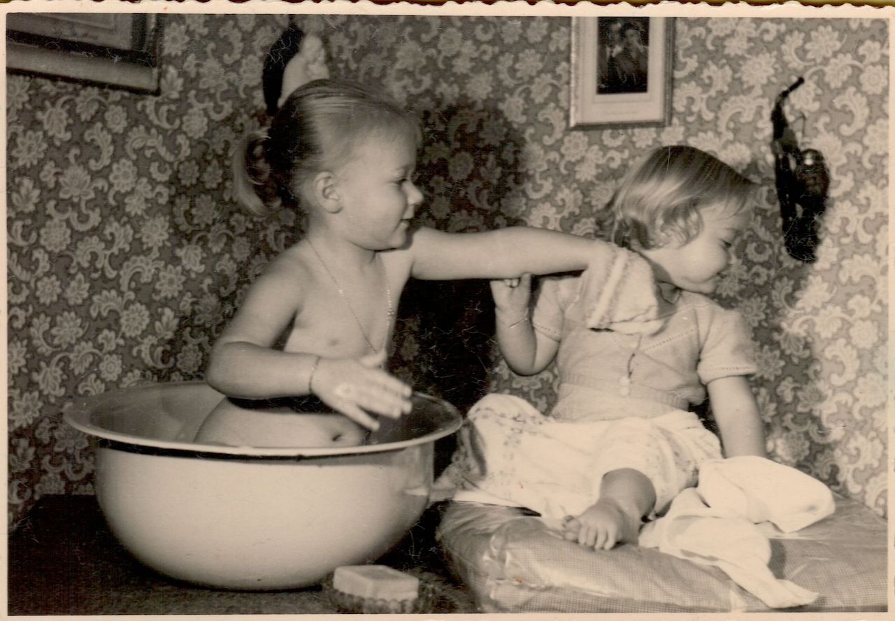 A young child sits in a large basin, playfully washing another child's face with a cloth.