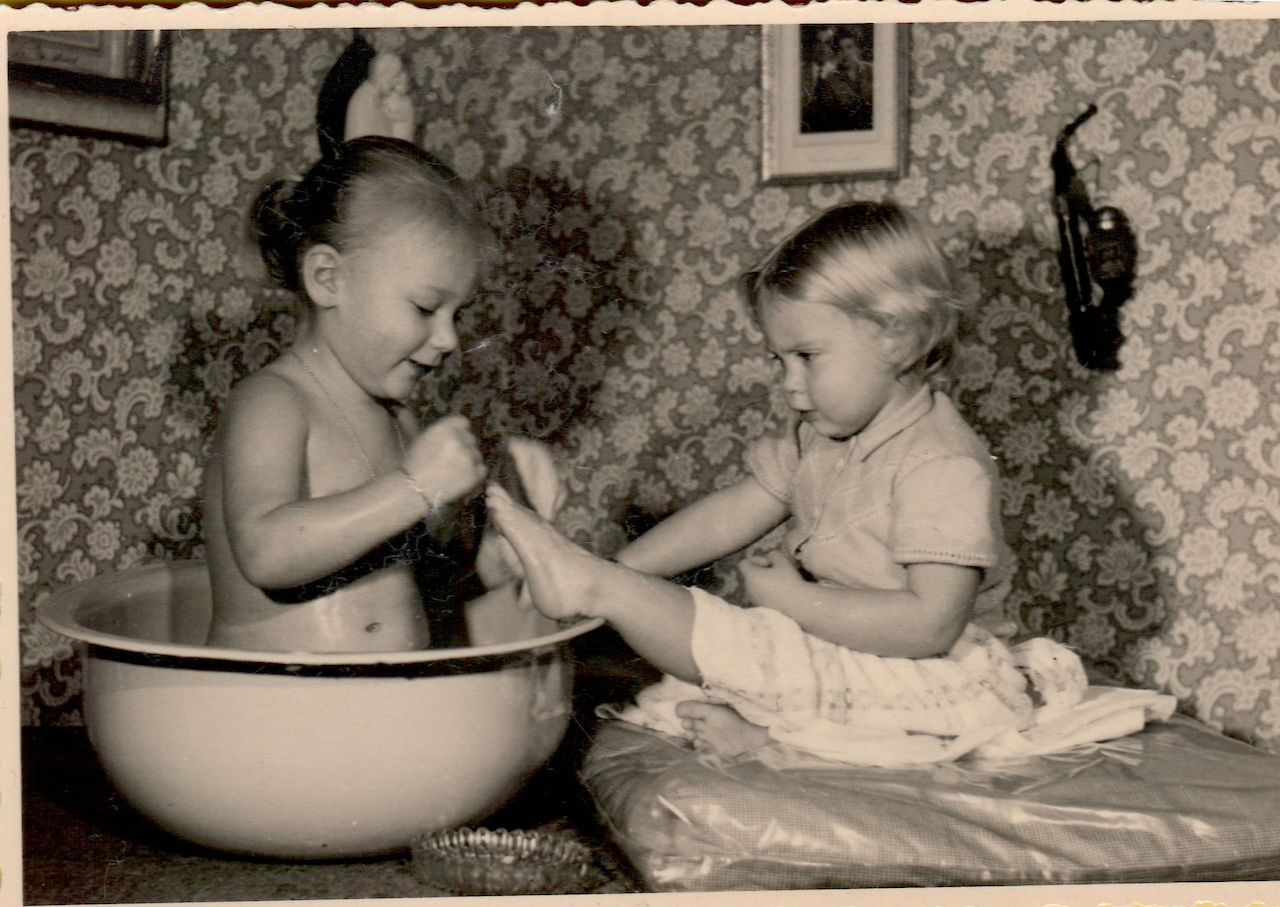 A young child sits in a large basin, playfully washing another child's feet in a cozy indoor setting.