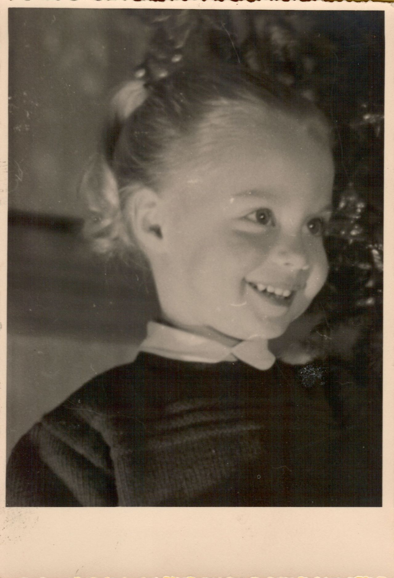 A young child with a bright smile looks off to the side, wearing a dark sweater with a white collar.