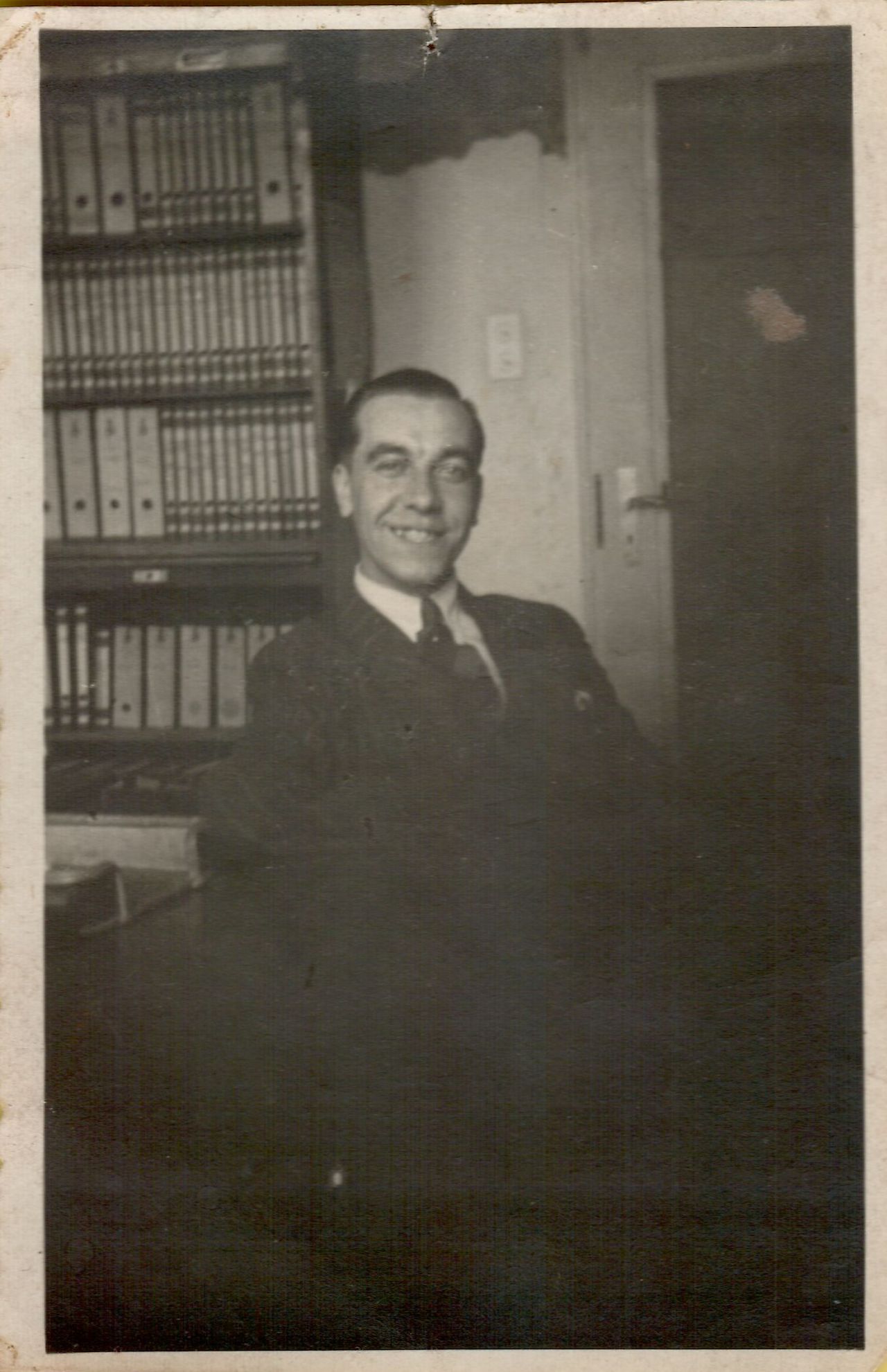 A man in a suit sits and smiles in an office with shelves full of binders behind him.