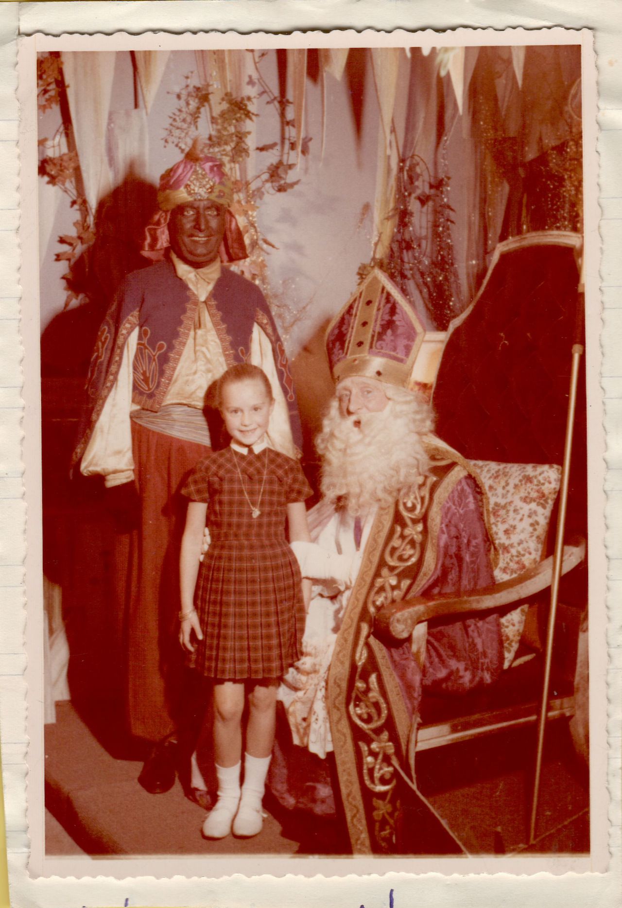 A young girl stands next to Sinterklaas and another costumed figure, smiling for a festive holiday photo.