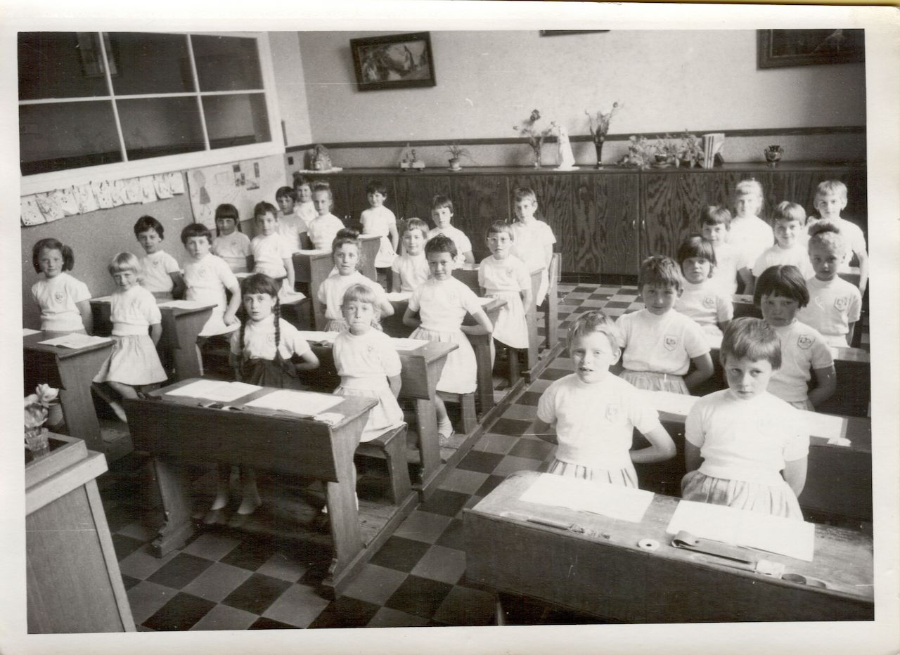 A classroom of young students in uniform stand at their desks, appearing to greet someone or respond to a teacher.