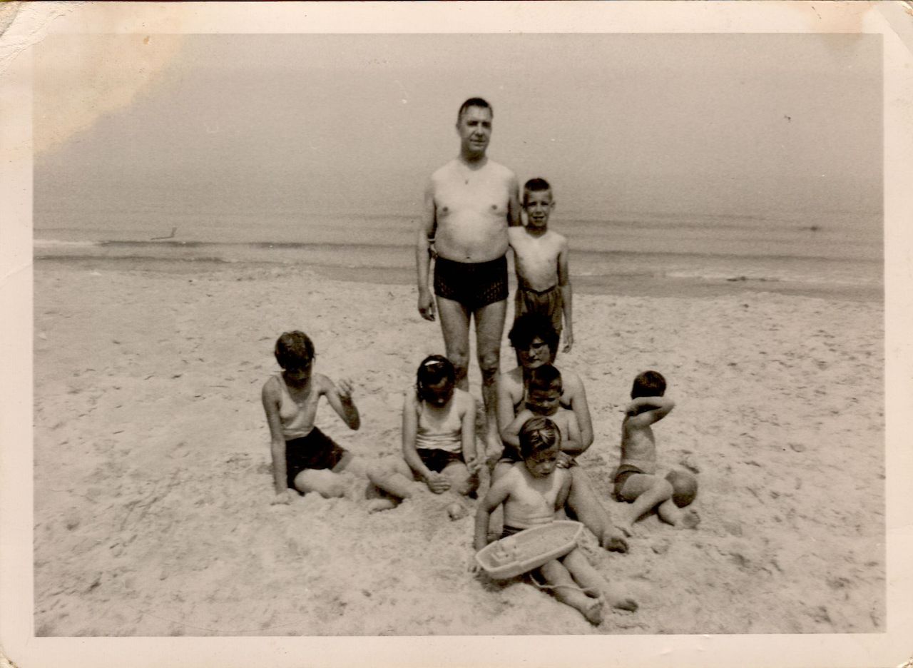 A man stands on a sandy beach with children sitting and playing around him, some holding toys.
