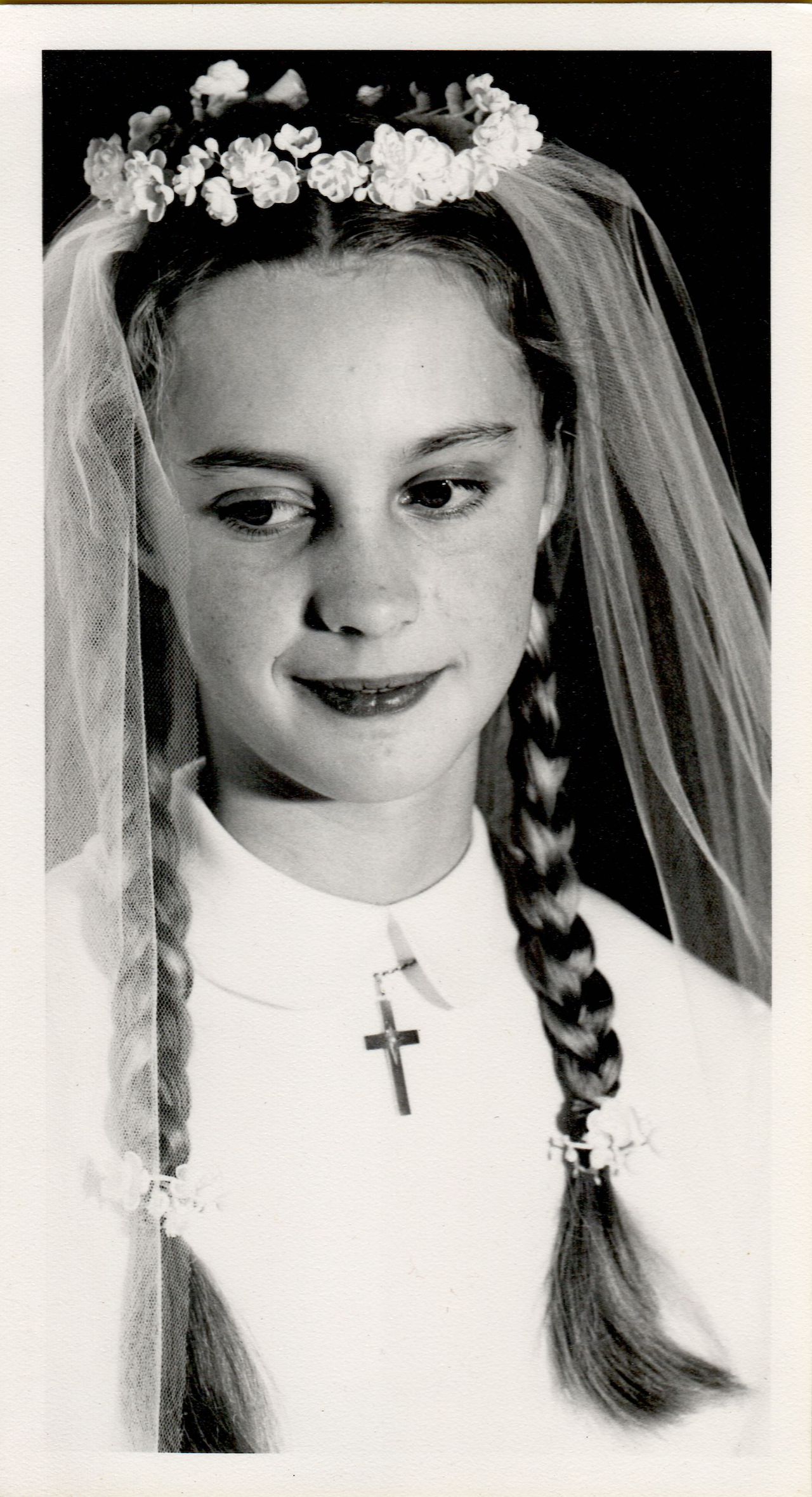 A young girl in a white dress with a veil and floral headpiece, wearing a cross necklace, looks downward.