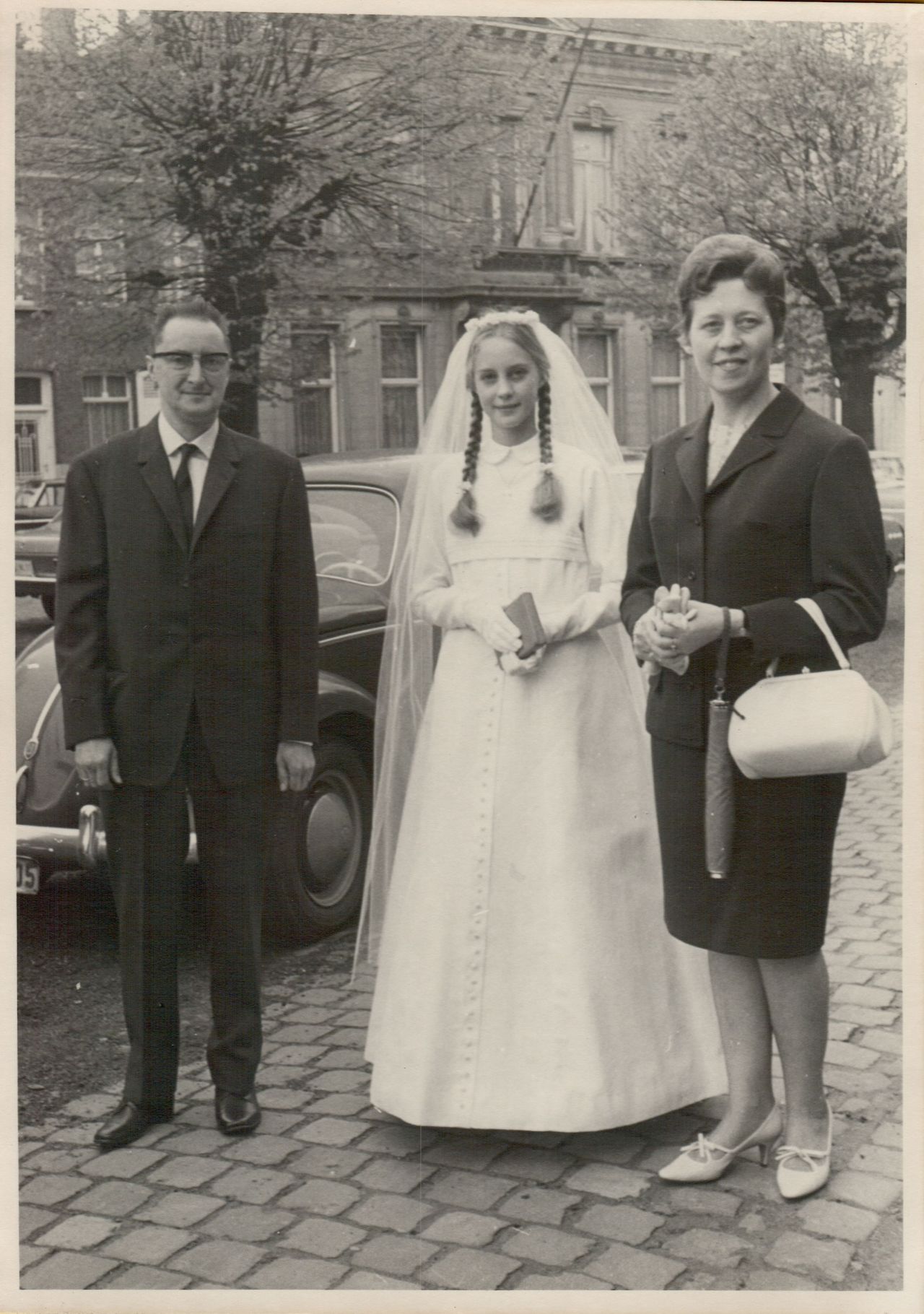 A young girl in a white dress stands between two adults, likely celebrating her First Communion.