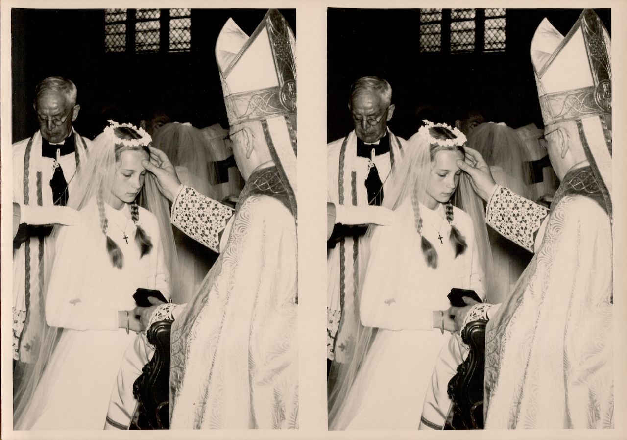 A young girl in a white dress and veil receives a religious blessing from a bishop during communion.