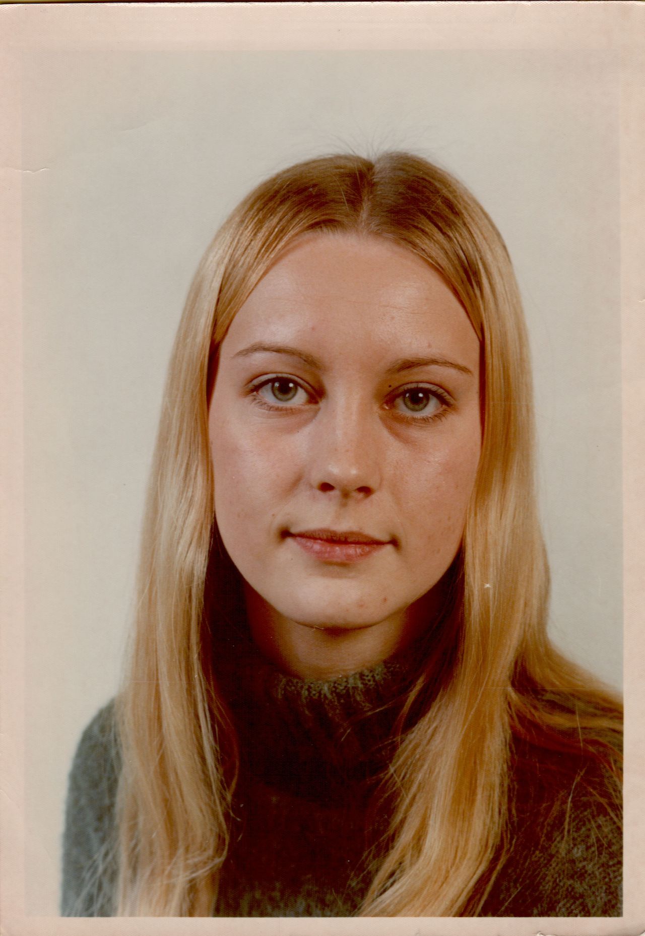 A woman with long blonde hair wears a dark sweater and looks directly at the camera with a calm expression.