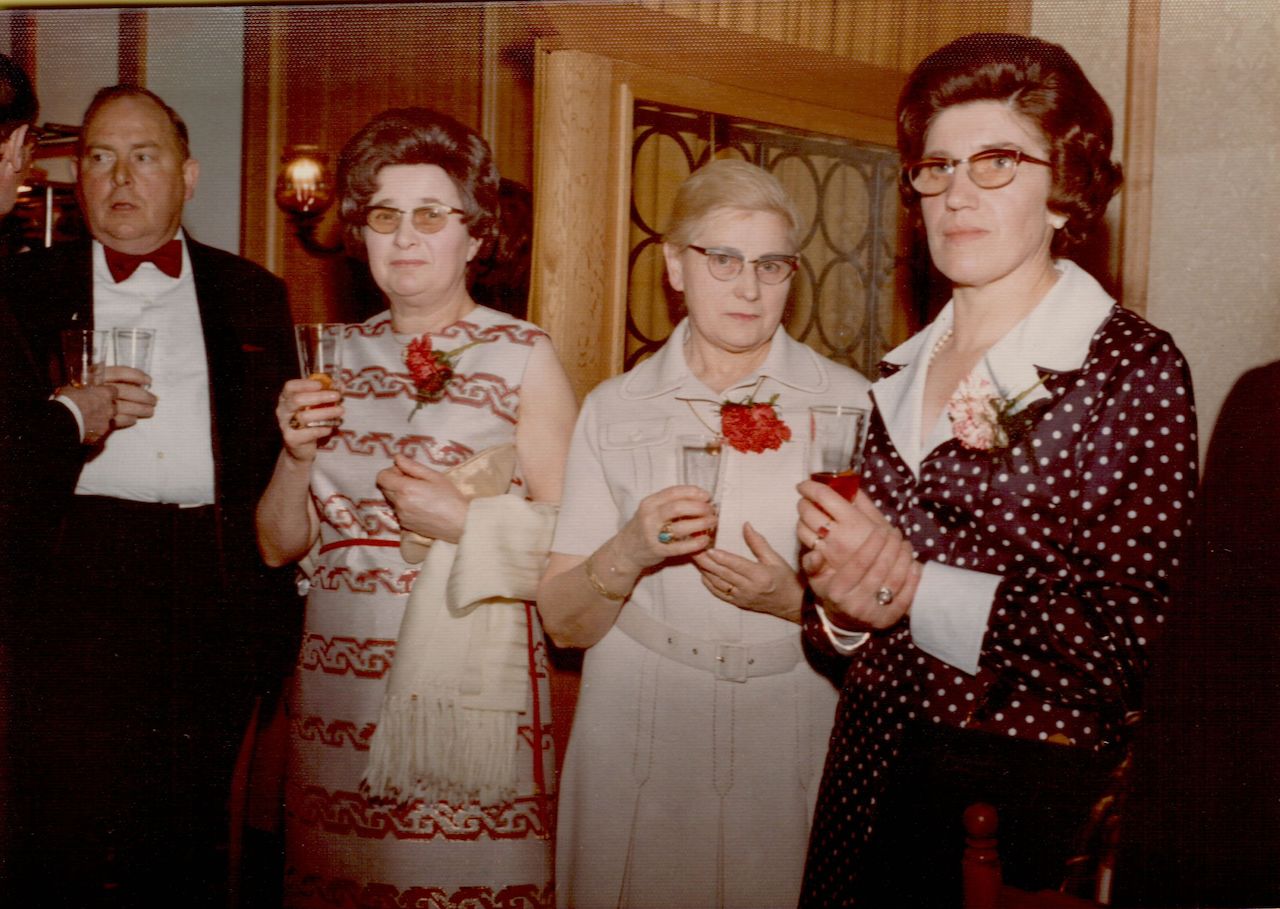Four adults dressed formally stand together at an indoor event, holding drinks and wearing red carnation boutonnieres.
