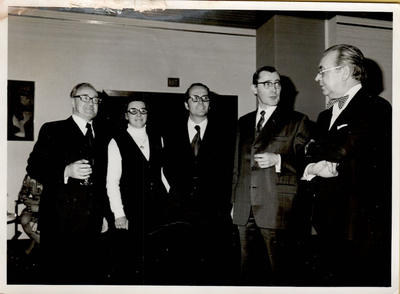 Five people in formal attire stand together indoors, engaged in conversation.