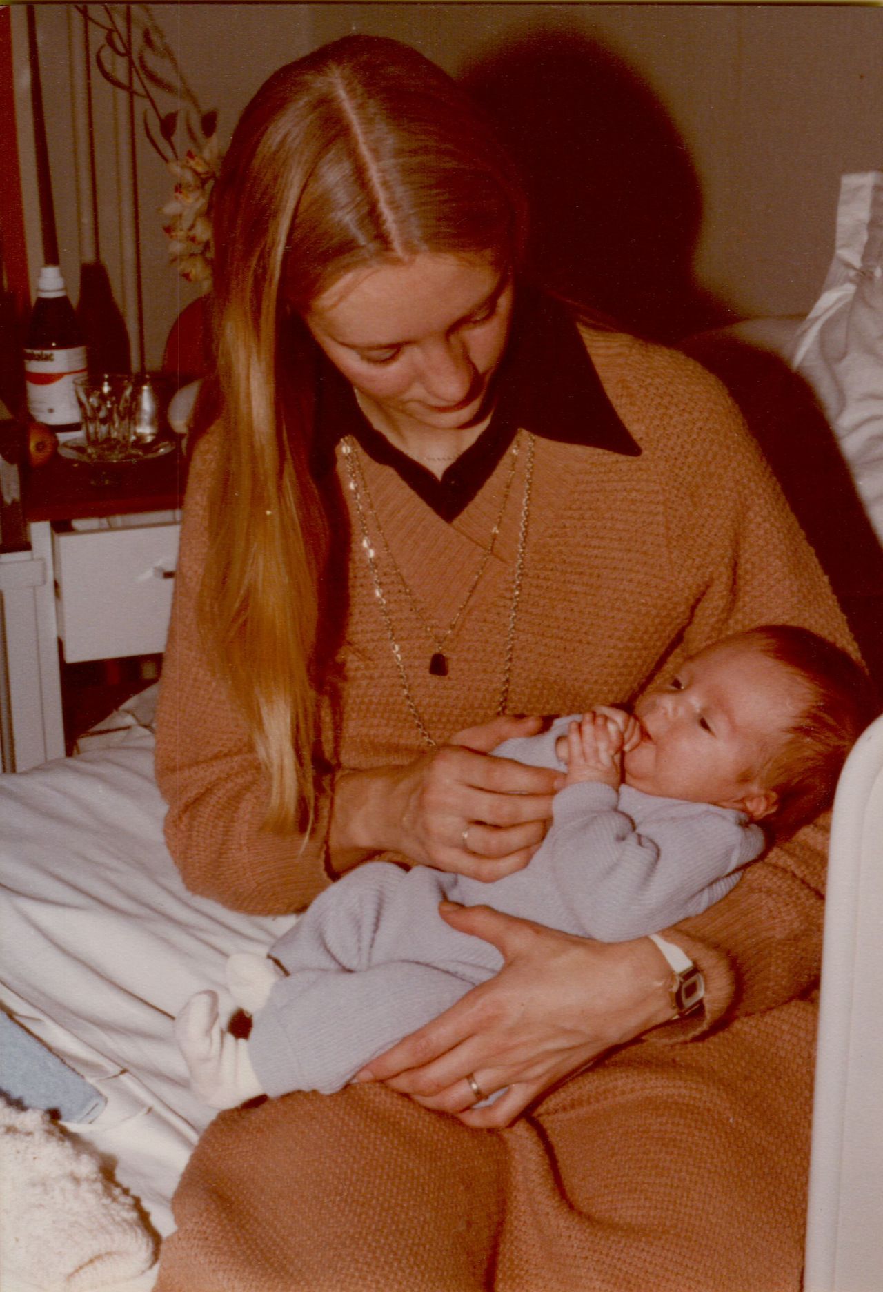 A woman sits on a bed, gently holding a newborn baby dressed in light blue, who is sucking their fingers.