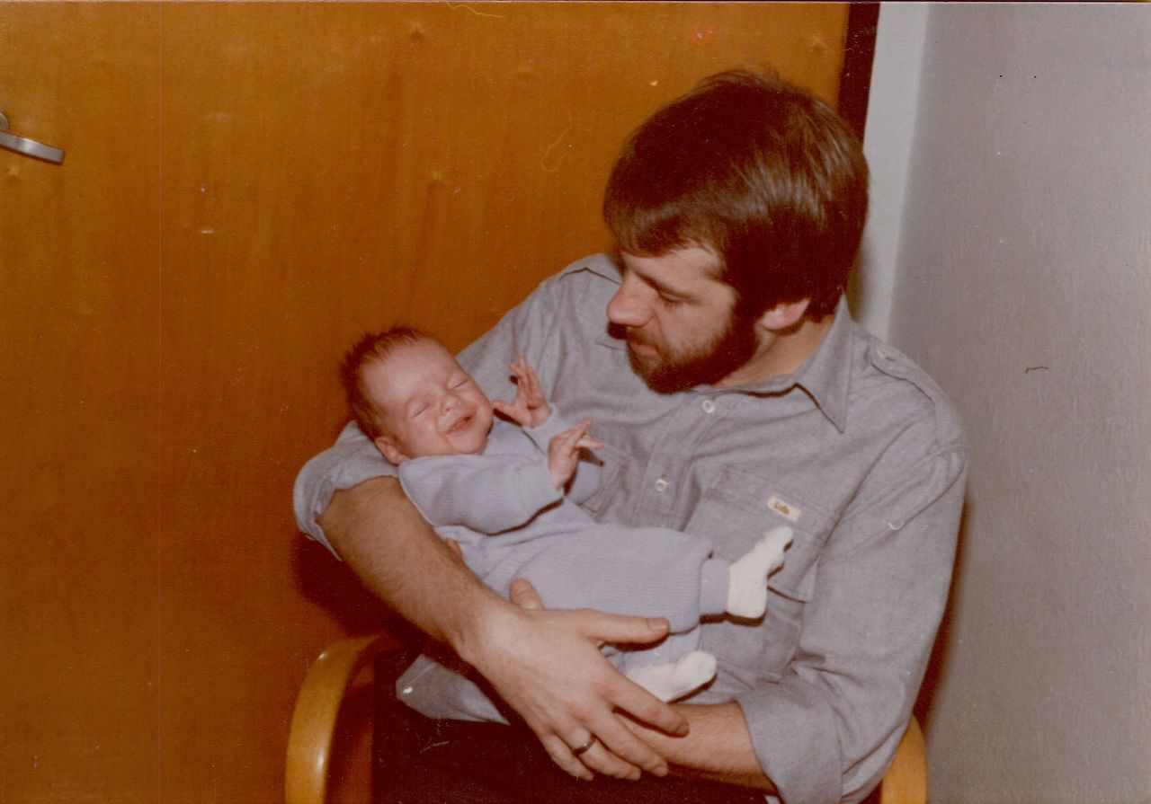 A man in a gray shirt holds a newborn baby dressed in light blue, who appears to be smiling or squinting.