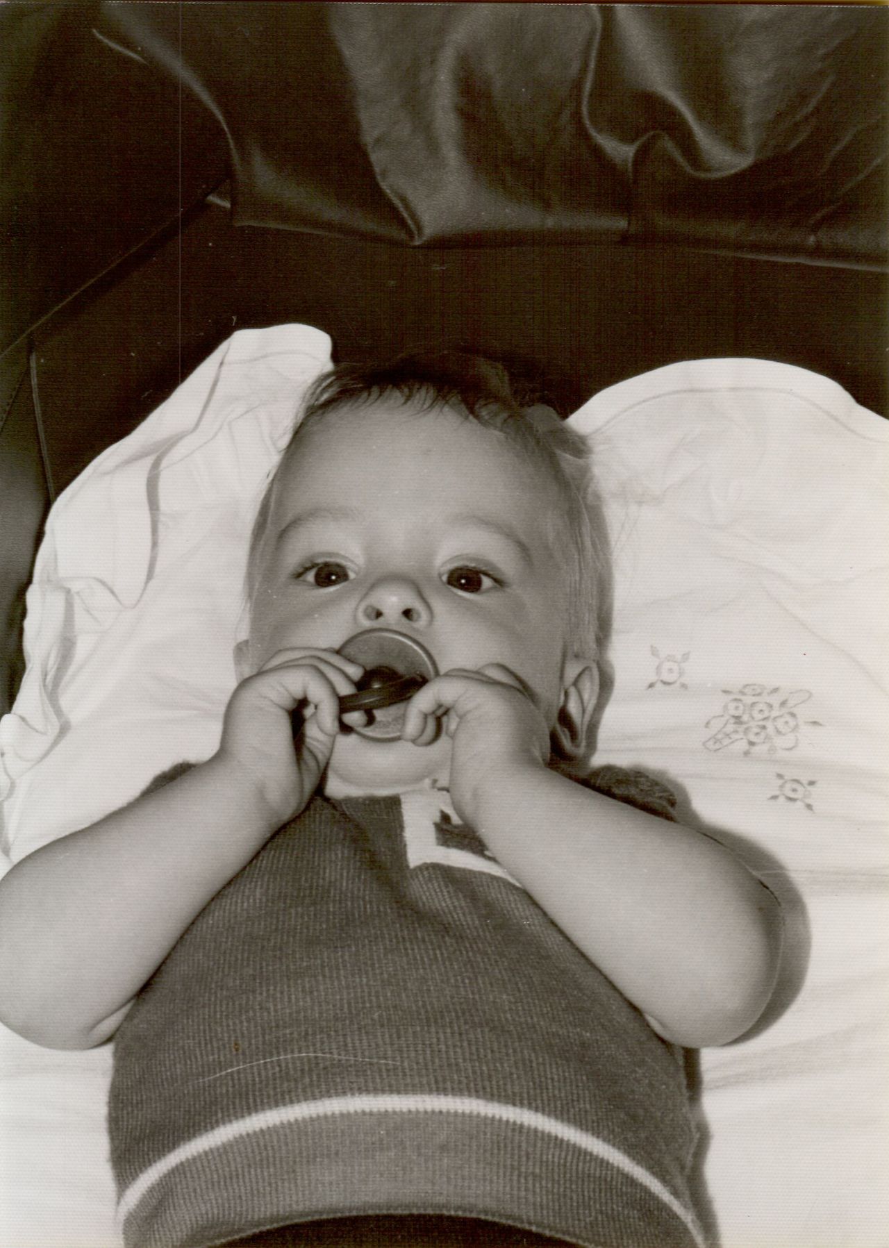 A baby lies on a pillow, holding and sucking on a pacifier while looking up.