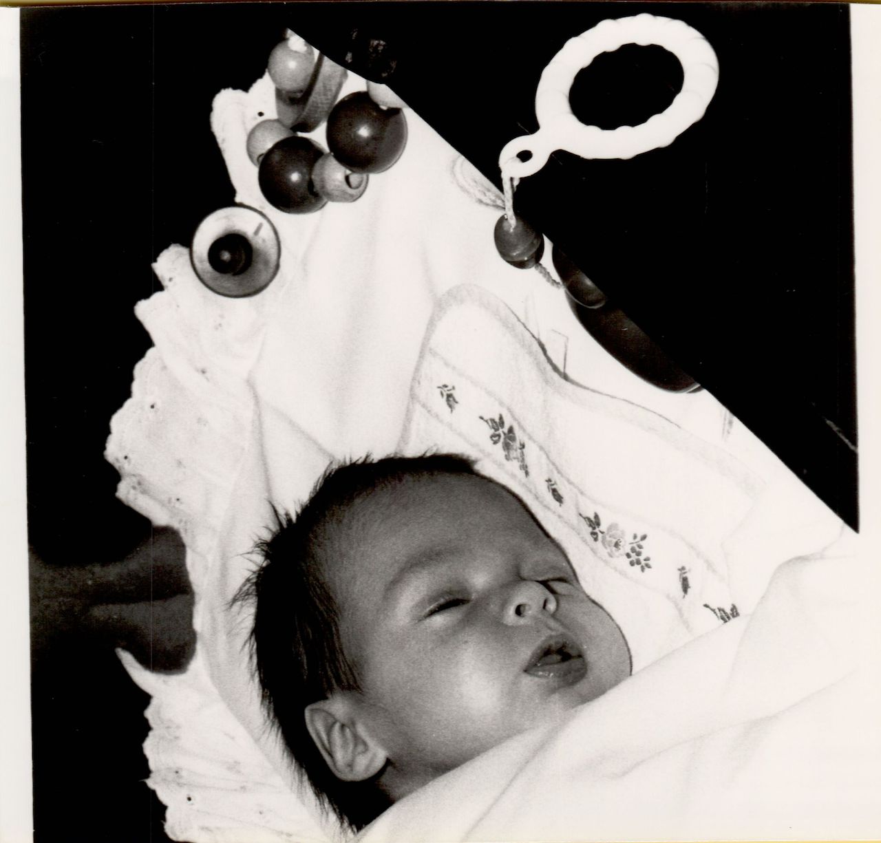 A baby lies swaddled in a blanket, eyes slightly open, with a hanging toy visible above.