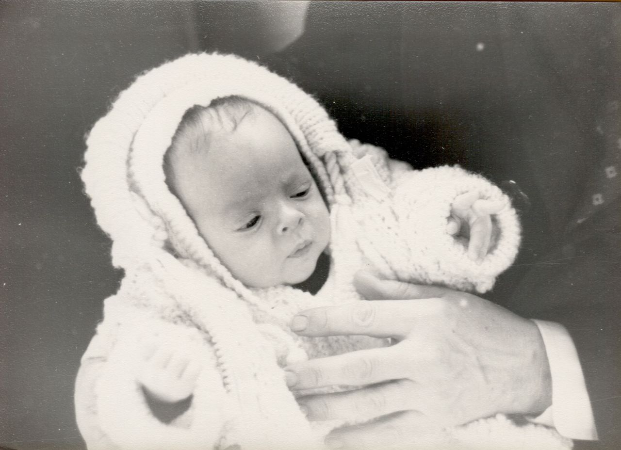 A baby dressed in a knitted outfit is held by an adult during a baptism ceremony.