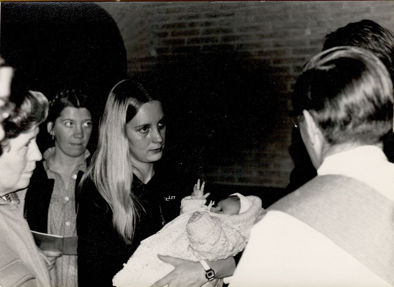 A woman holds a baby dressed in a knitted outfit during a baptism ceremony, surrounded by onlookers and a clergyman.
