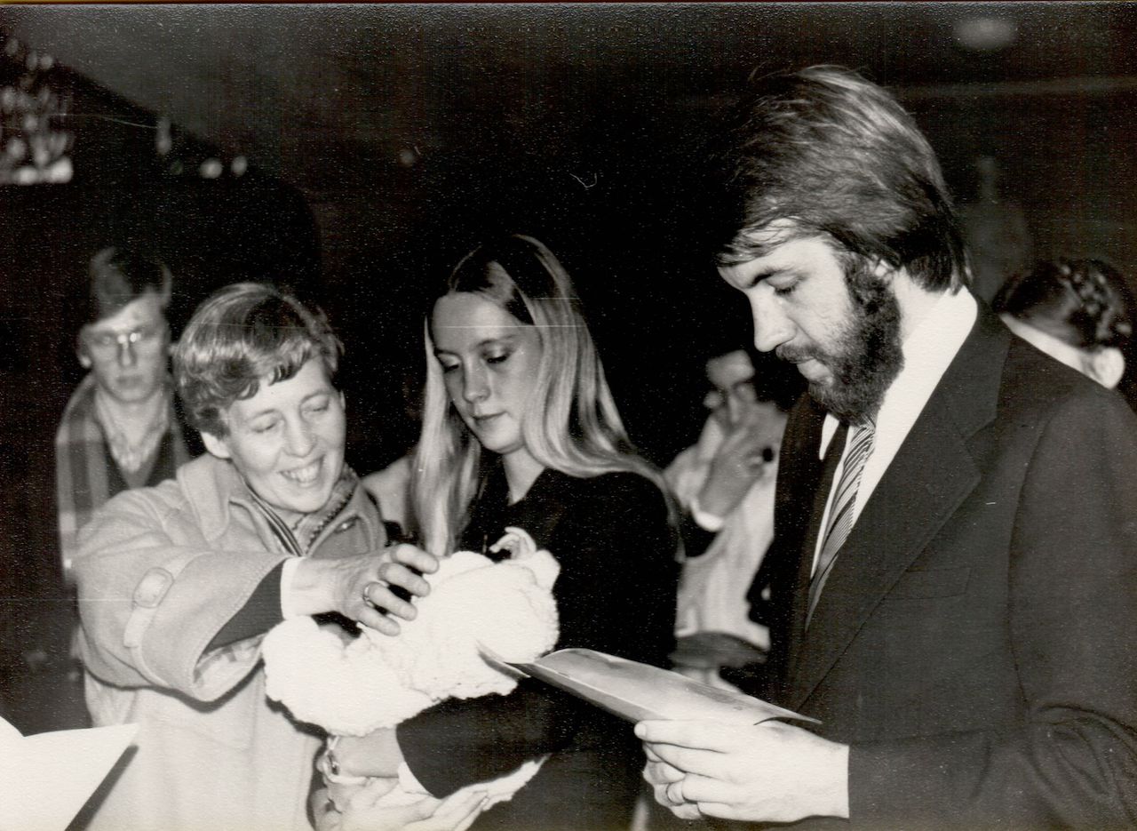 A woman smiles while touching a baby in a baptism ceremony, as a man reads from a paper.