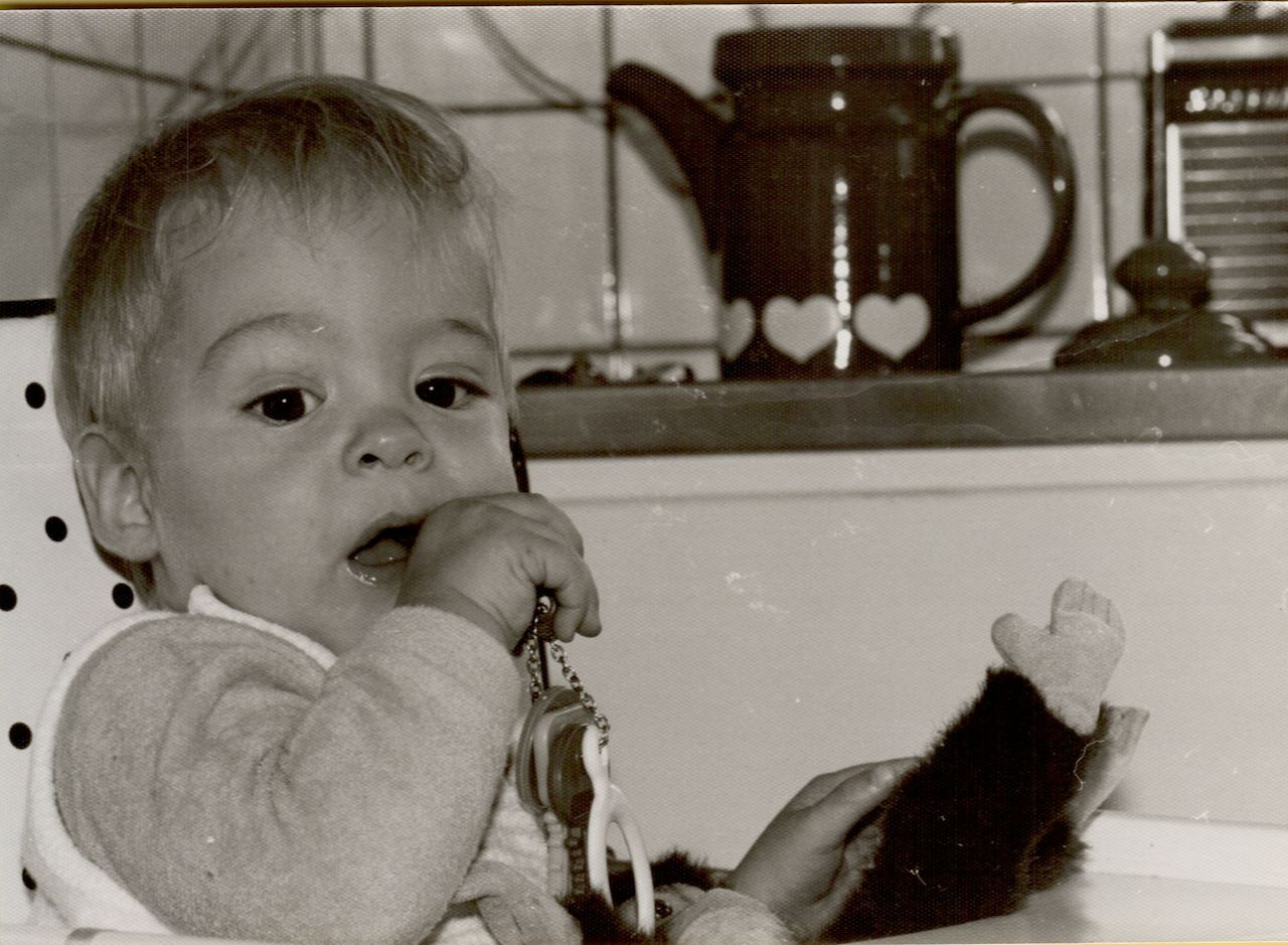 A young child sits in a high chair, chewing on a pacifier clip while holding a stuffed toy.
