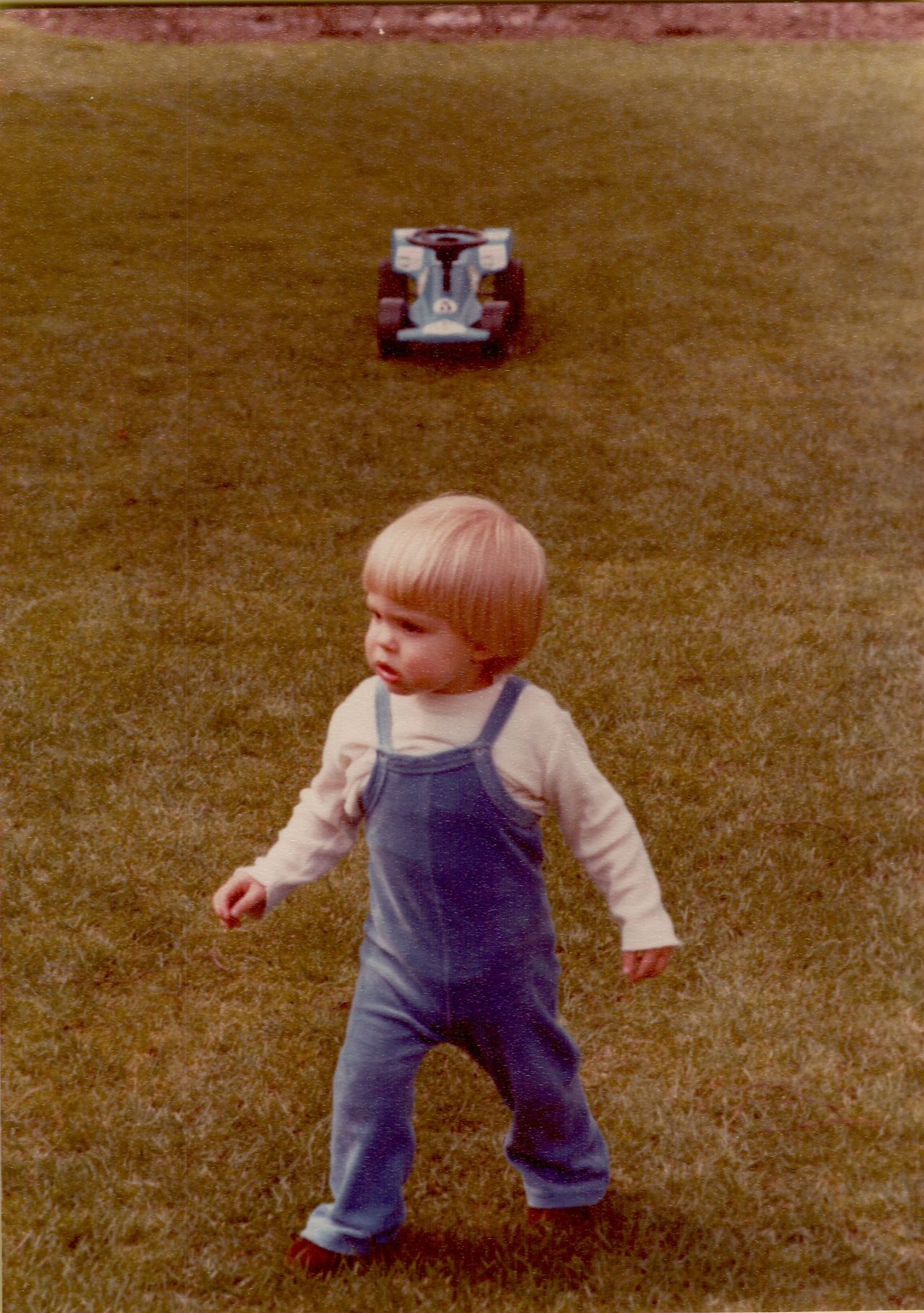 A young child in blue overalls walks on grass, with a small toy race car behind them.