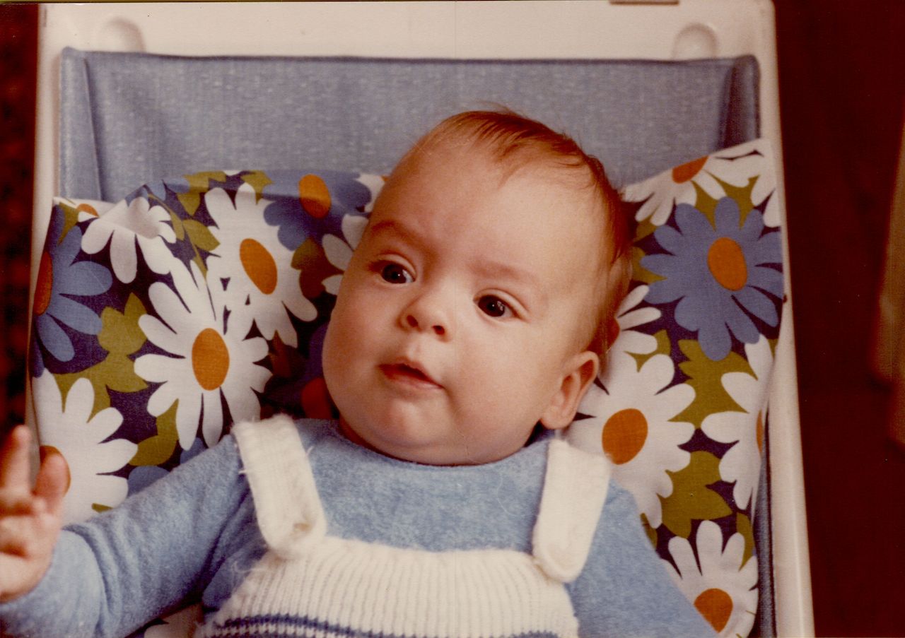 A baby sits in a chair with a floral cushion, wearing a blue sweater and white overalls.