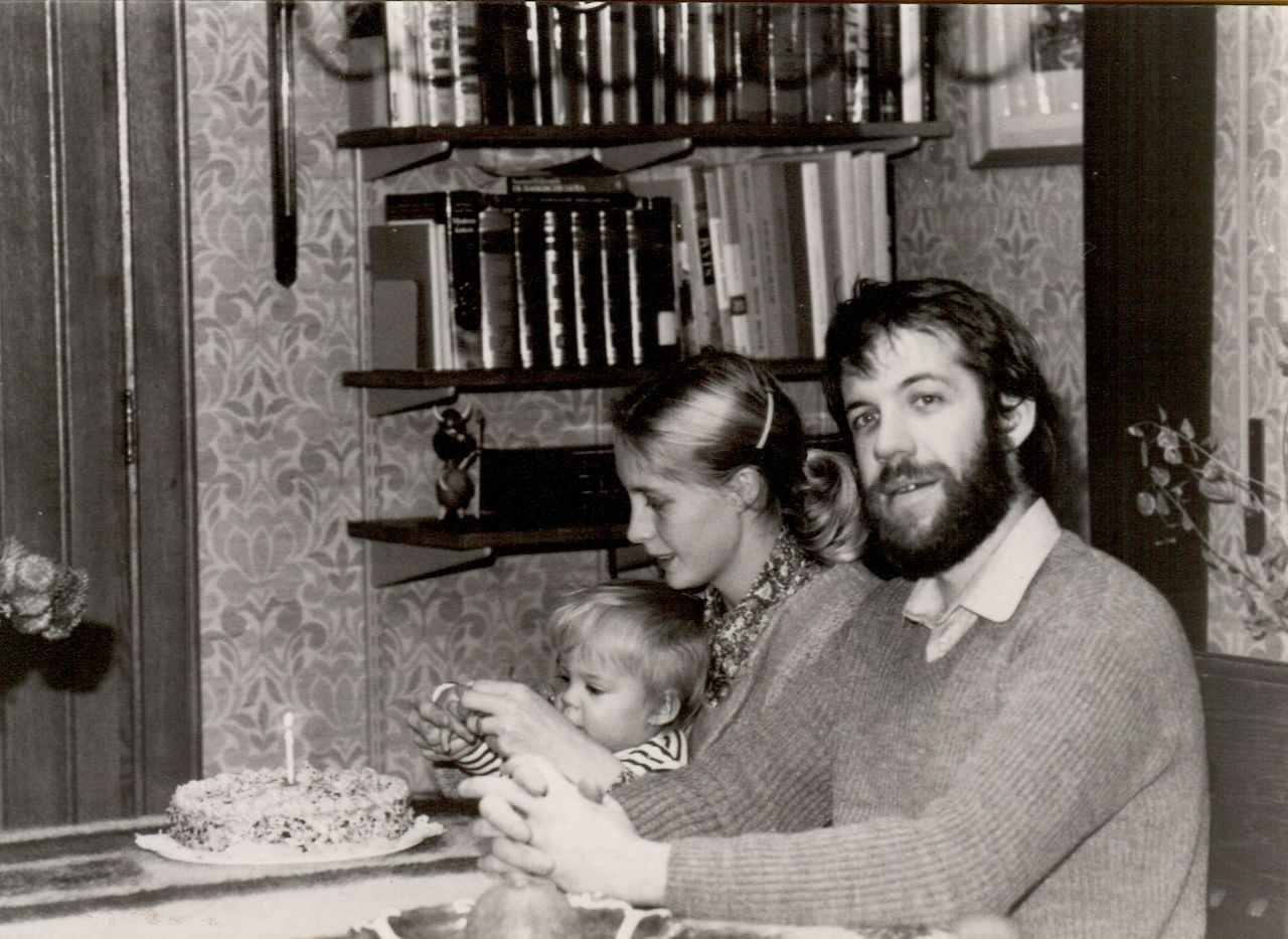 A baby sits with two adults at a table, looking at a small birthday cake with one candle.