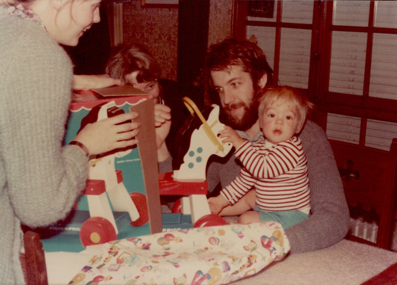 A baby in striped clothes sits on a man's lap, holding a toy horse while others open a box.