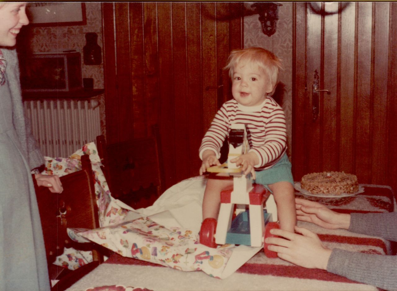 A one-year-old child sits on a toy horse, surrounded by family, with a birthday cake nearby.