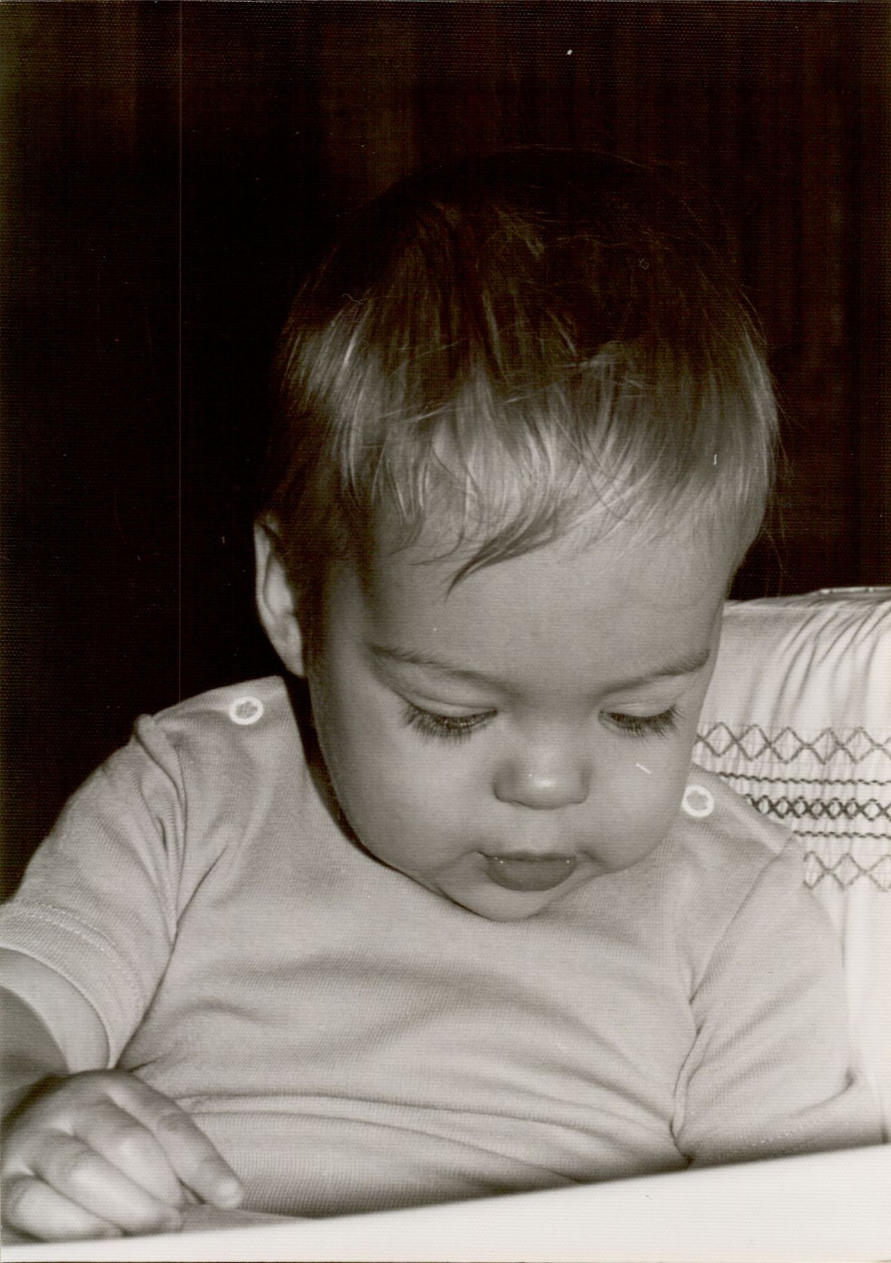 A baby sits upright, looking down with a focused expression, wearing a short-sleeved shirt.