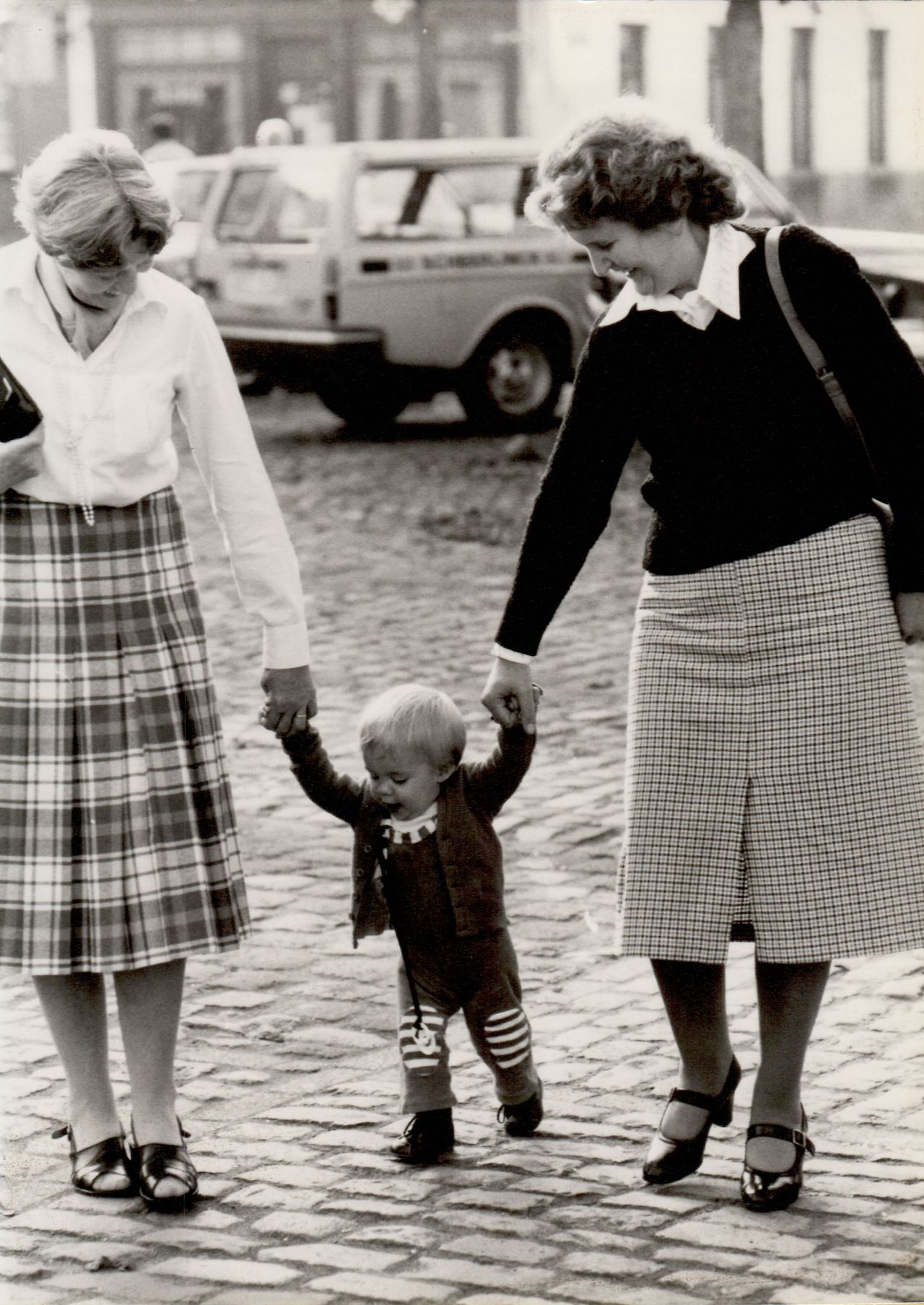 A young child walks on a cobblestone street, holding hands with two women for support.