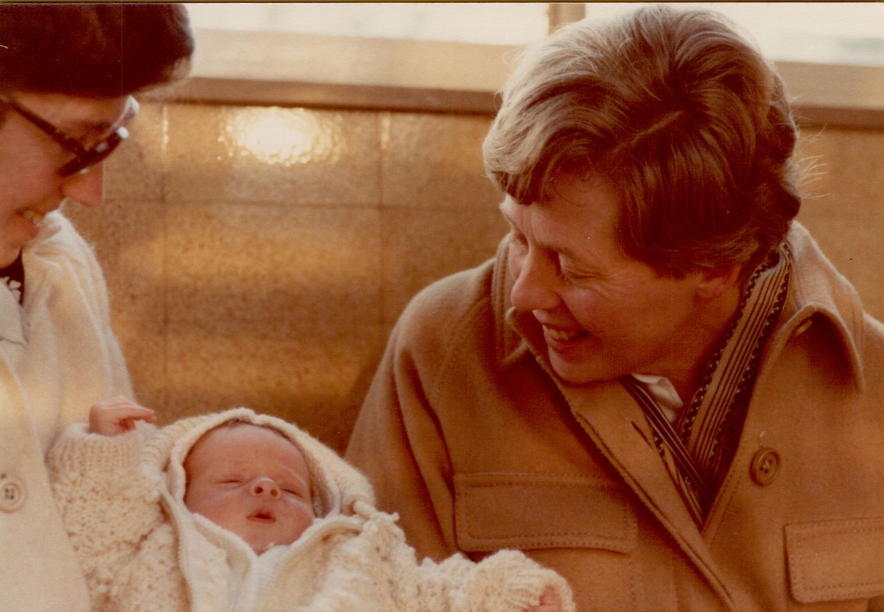 A baby in a knitted outfit is held by two smiling women, who appear to be grandmothers.