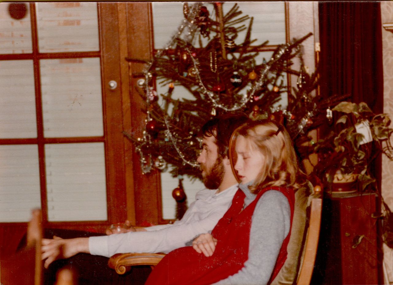 A pregnant woman in a red dress sits next to a man in a chair near a decorated Christmas tree.