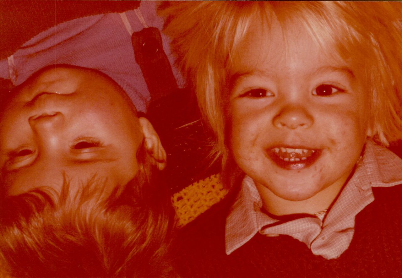 Two young children lie close together, one smiling brightly while the other rests with eyes partially closed.