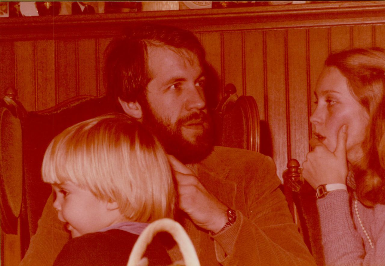 A man with a beard sits with a young child on his lap while a woman listens thoughtfully nearby.