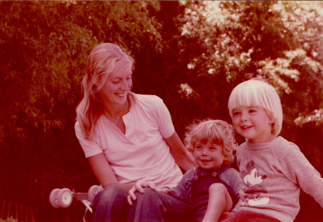 A woman sits outdoors, smiling at two young children who are also smiling.