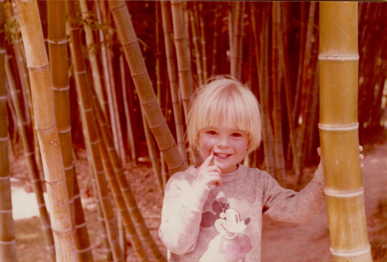 A young child with blonde hair smiles, touching their face, while holding onto a bamboo stalk in a forest.