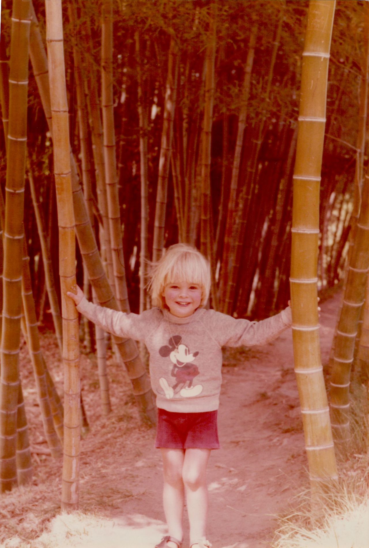 A smiling child stands on a dirt path, holding onto two bamboo stalks with both hands.