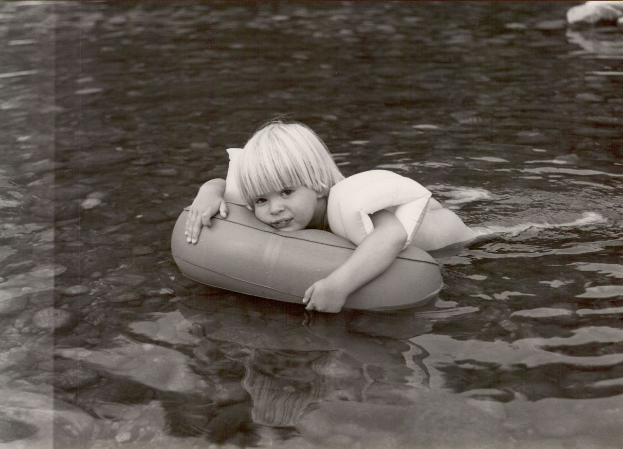 A young child with floaties holds onto an inflatable ring while floating in a shallow river.