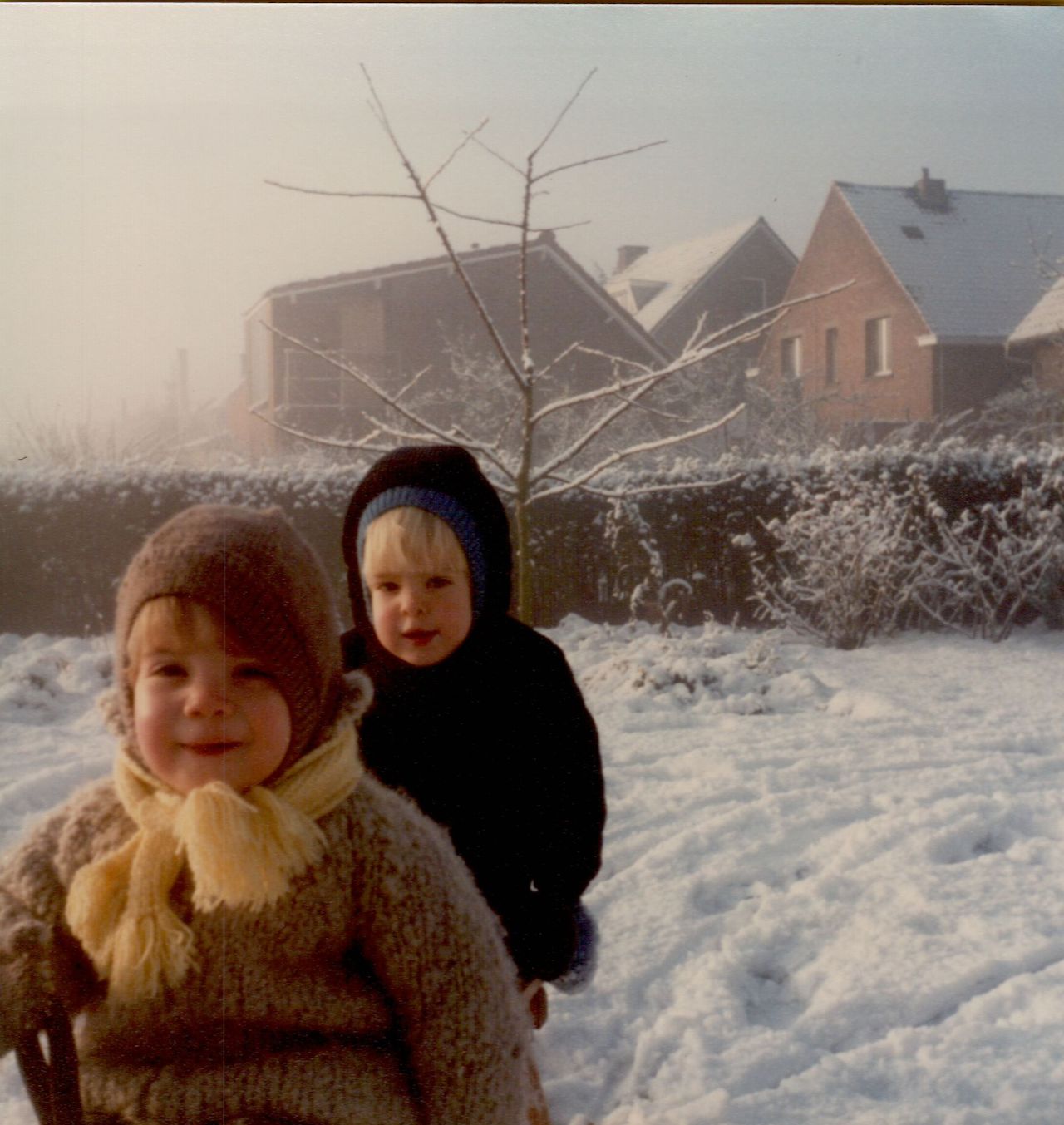 Two children in warm winter clothes play in the snow, one sitting on a sled while the other stands behind.