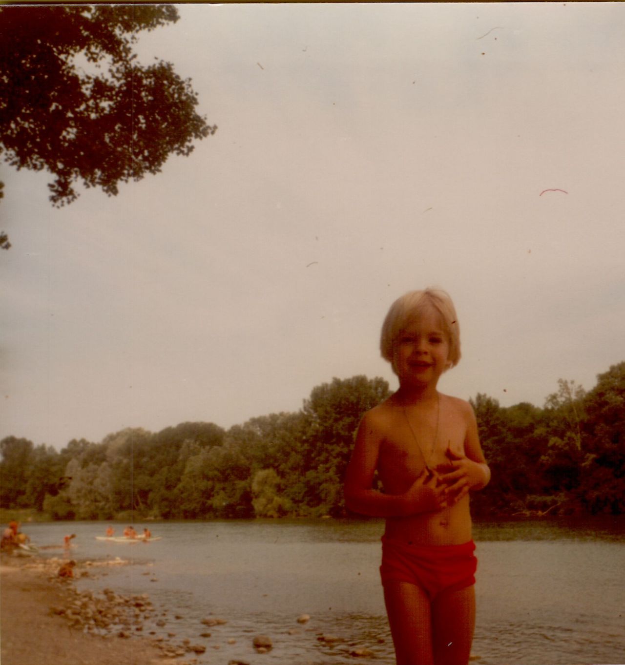 A young child in red swim trunks stands near a river, smiling at the camera.
