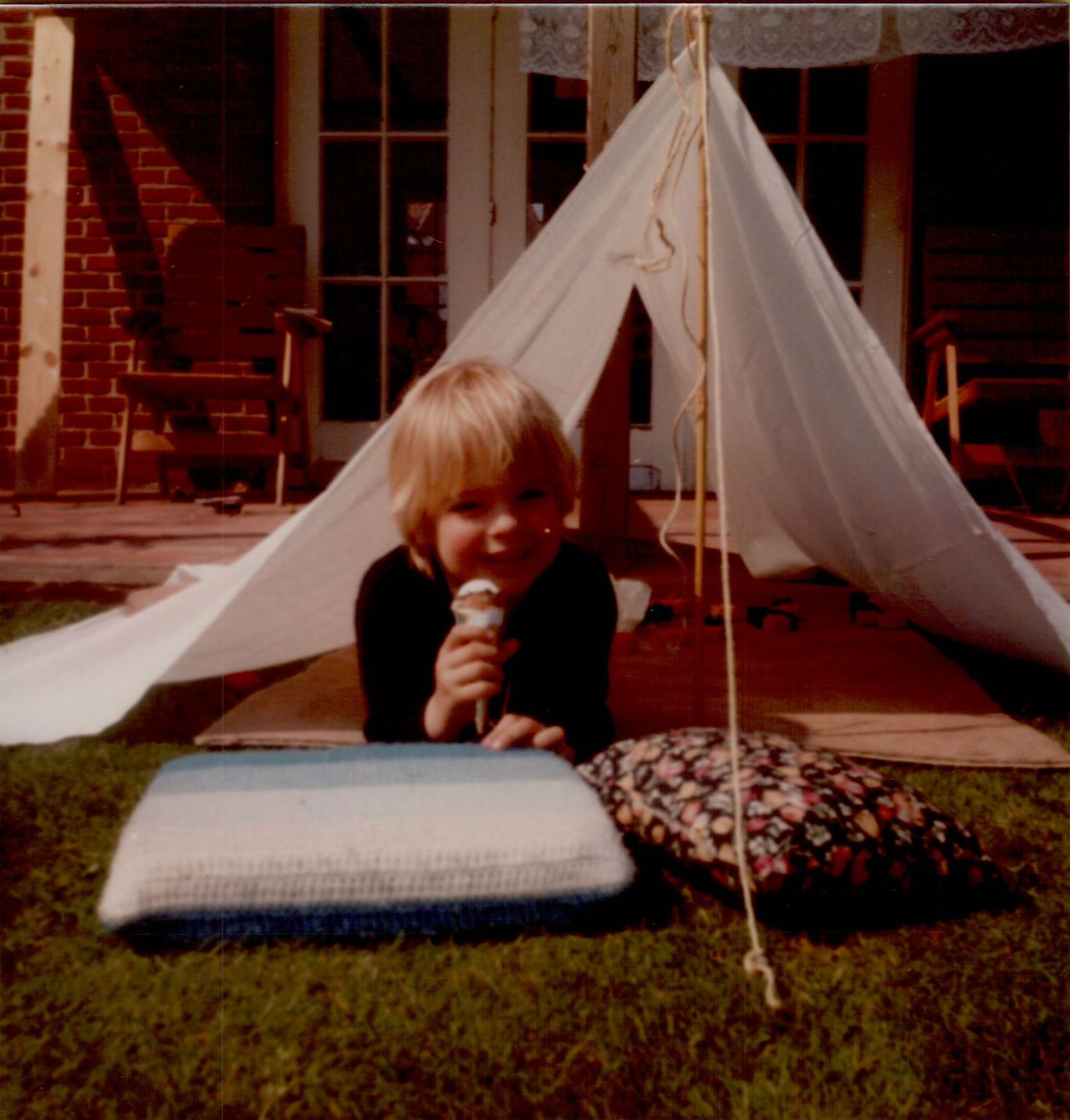 A child lies on the grass, holding an ice cream cone in front of a small homemade tent.