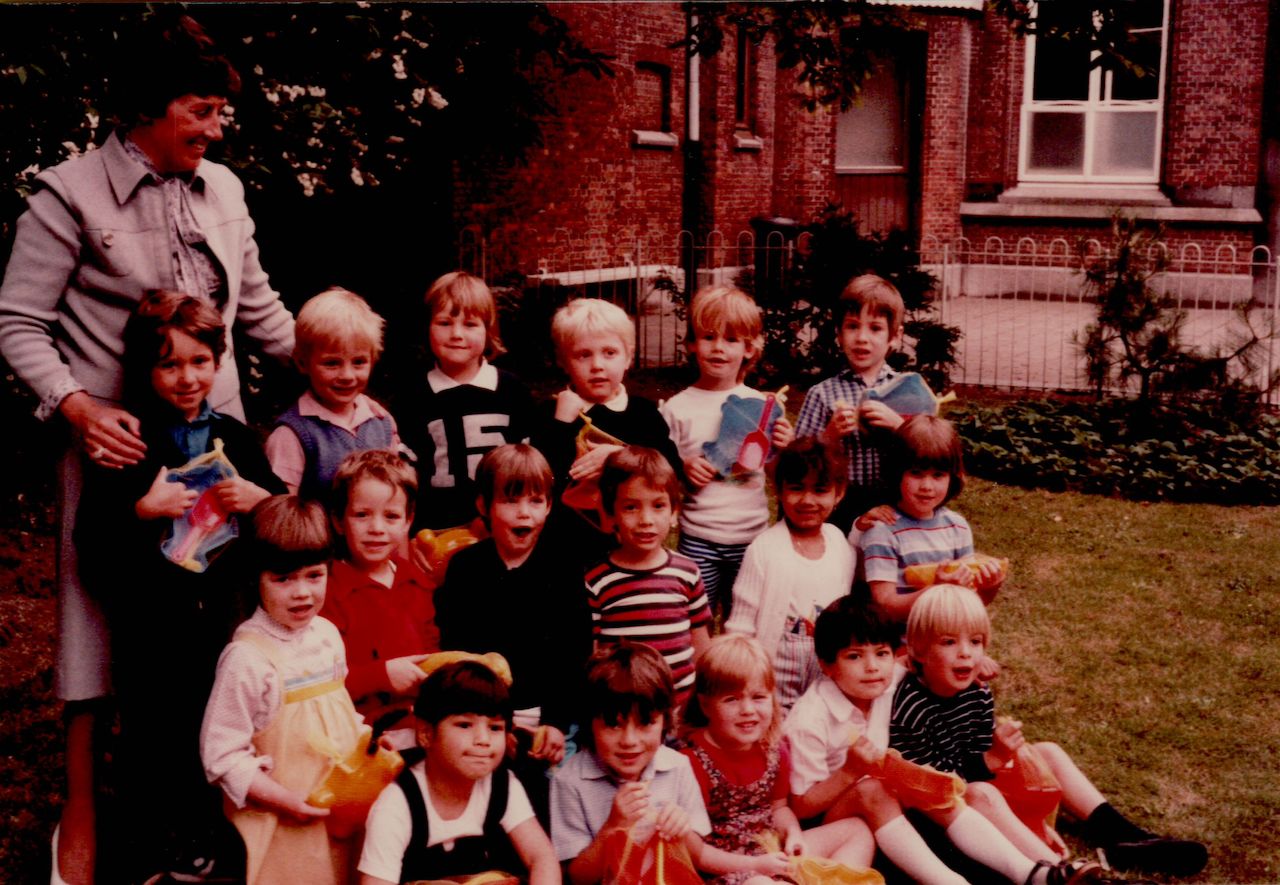 A group of young children and their teacher pose for a class photo outdoors, holding small colorful bags.