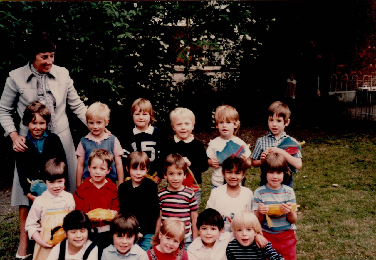 A group of young children and their teacher pose for a class photo outdoors, some holding small toys or crafts.