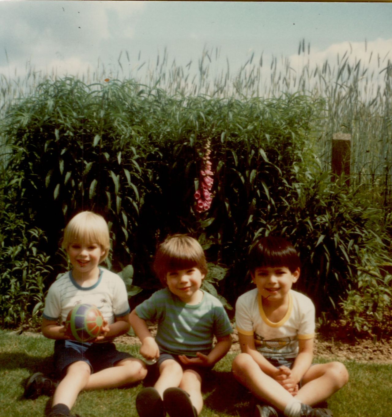 Three young children sit on the grass, smiling at the camera.